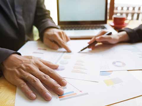 two-workers-drawing-on-stack-of-papers-with-graphs-and-computer-in-background